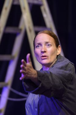 Eine Schauspielerin steht vor einer Leiter und streckt den Arm nach vorn.